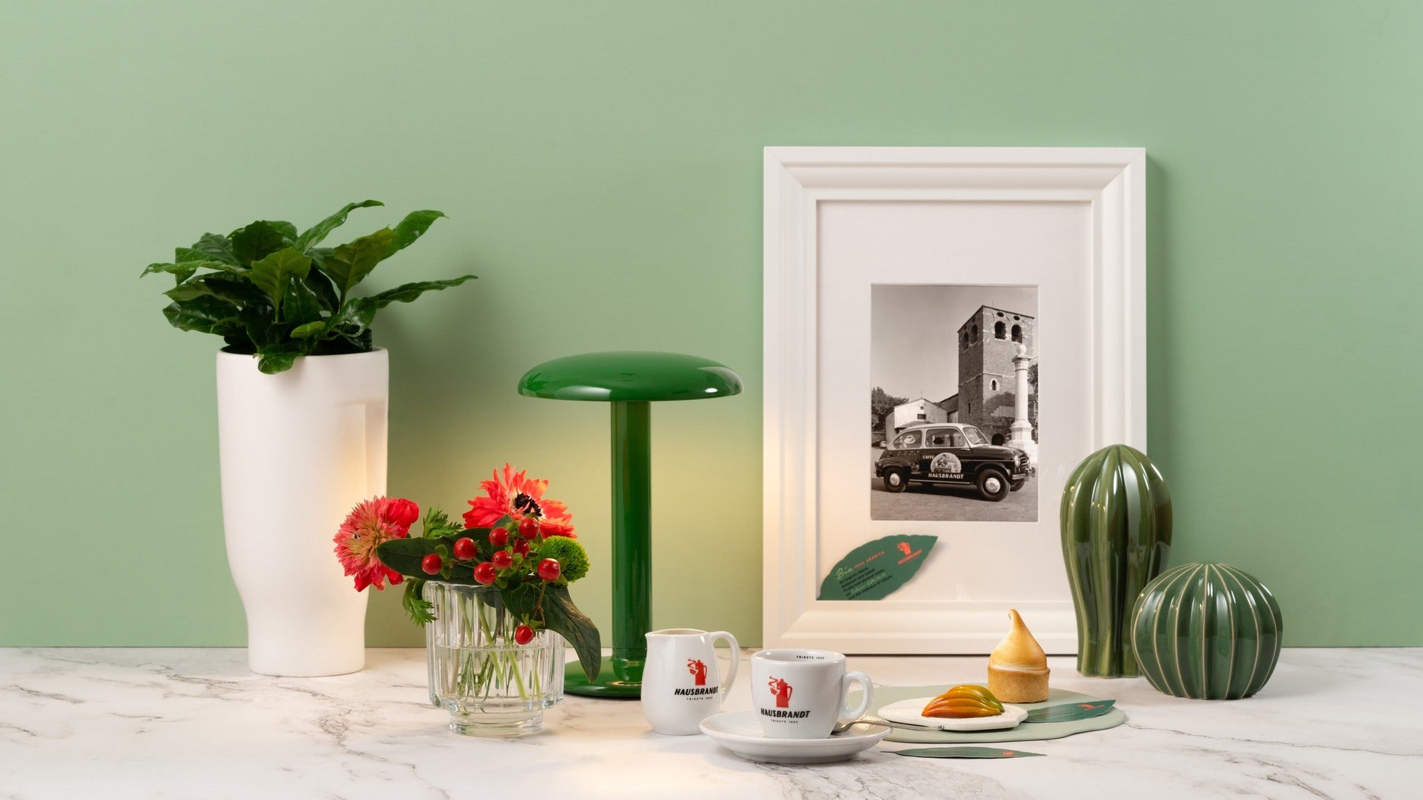Decorative table setting with a lamp, plants, and framed picture against a green wall.
